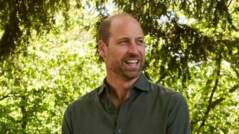 Prince William smiles broadly as he stands in front of trees during an event. He is wearing a dark green shirt.