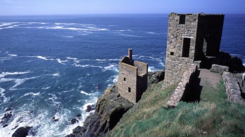 This picture shows the iconic engine houses of the historic Botallack Mine on the rugged Cornish coastline. The structures are perched dramatically on cliffs overlooking the Atlantic Ocean, with waves crashing against the rocks below. These stone buildings were part of Cornwall’s tin and copper mining industry during the 19th century and are now preserved as part of the UNESCO World Heritage Site for Cornish Mining. The scene captures the wild beauty of the coast, with deep blue sea, clear skies, and the stark remains of industrial heritage blending into the natural landscape.
