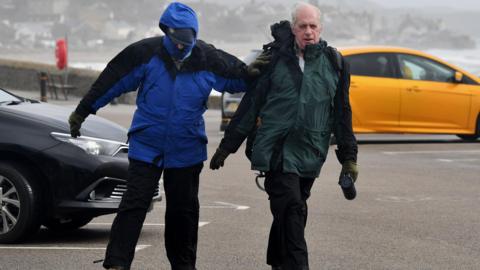 Two men are walking across a car park in the wind wearing anoraks. One of them looks like he is almost being blown off his feet. There is the sea behind them and a black and a yellow car are parked in the car park.