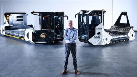 A man standing an a dark warehouse that has dim lighting. He is standing with arms crossed in front of two self-driving vehicles that are used to move airport baggage. He is wearing suit trousers and a blue shirt and has short grey hair. He is smiling. 