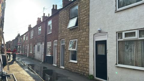Firefighters have taped off a section of a street with terraced houses on. There is water in pools in the street.