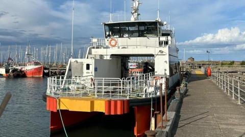 The Dart Fisher moored at a harbour. It is mostly white with yellow and orange paintwork at one end