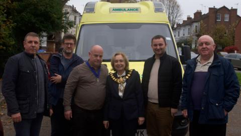 Standing in front of a decomissioned lime green ambulance are six people. They include the mayor of Leamington Spa in the centre with her chain of office. Next to her is her consort with his chain of office. Four men are stood either side of them. They are wearing dark coats.