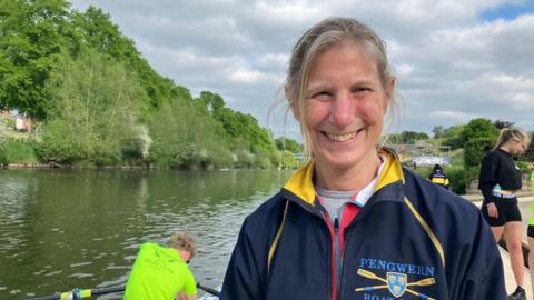 A woman with grey hair and a blue zip-up top with the word Pengwern on it stood in front of a river with a few people getting into boats behind her