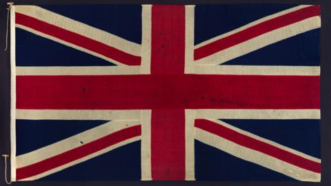 A Union Flag in a frame. It has a number of small holes in the fabric.
