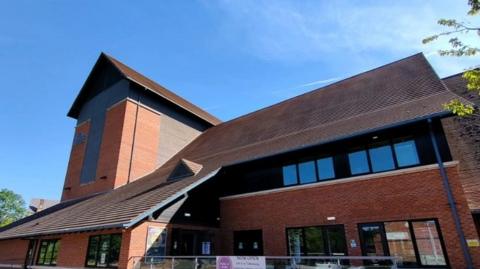 A red brick building with lots of glass windows. It is at least three storeys high. There are blue skies and some white cloud.