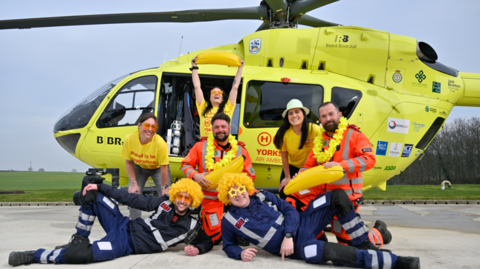 Seven members of the Yorkshire Air Ambulance team stand in front of a yellow air ambulance. Some are dressed in yellow t shirts, while others wear yellow sunglasses and wigs.