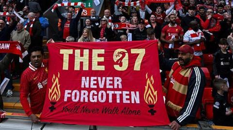 Liverpool fans at a football game hold a flag which reads 'The 97 Never Forgotten'.
