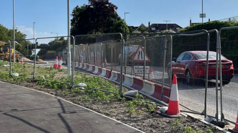 An image of a road. It is lined by traffic cones and metal fencing.