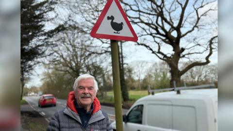 A man smiling straight at the camera and standing next to a road sign with a dodo on it.
