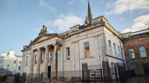 Bishop Street Court House is a large building, made of pale sandstone, with classical columns and sculptures. A large black gate surrounds the building. 