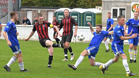 A man wearing a black and red football kit strikes a ball while footballers in a blue kit with white socks try to block his shot.