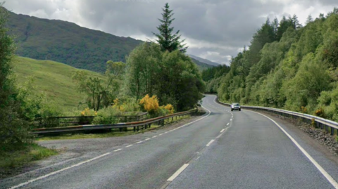 A winding single carriageway road with trees on each side and a car in the distance. There is a partially hidden road off the the left in the foreground, and a grassy hill in the distance.