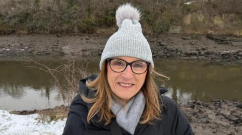 Allyson Timm standing by the edge of the River Wear. She has shoulder-length blonde hair and is wearing dark-rimmed glasses, a grey scarf and bobble hat. Snow can be seen behind her close to the river.