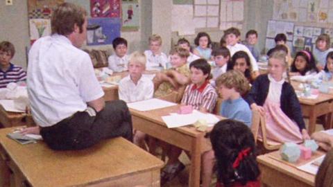 A classroom of children sit around a man on a teacher's desk.