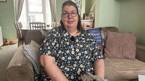 Nikki Paine pictured at her home in Ashford, Kent. She is sat on a beige sofa and has a dog at her feet. She is wearing a floral dress and has long brown hair and glasses.