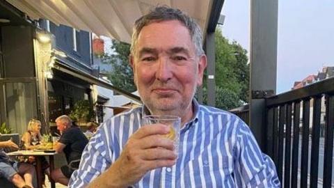 Aled, a man with black and grey hair wearing a blue and white striped shirt. He is smiling at the camera while holding a glass with a drink inside. He is sat in outdoor area of a bar/restaurant.