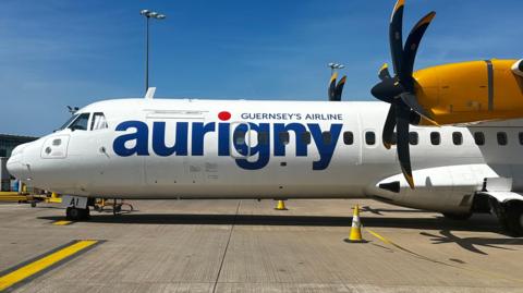 The side of a plane. It has Aurigny livery in blue colour text.