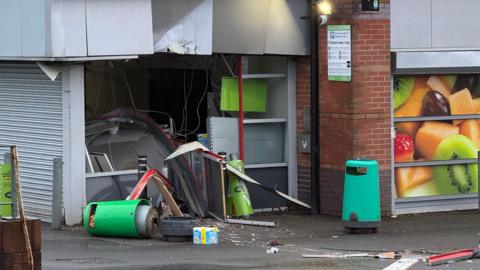 The front of a supermarket, with extensive damage to an entrance foyer. There is a green bin on its side and debris from the damage. There is another green bin and signage on a wall to the right hand side of the image.