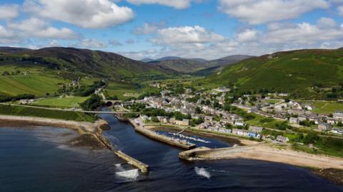 An aerial view of the coastal community of Helmsdale in the Highlands. Helmsdale is at the mouth of a river, and has a harbour and rows of white-walled houses. Green hills rise behind the village.