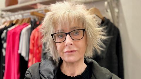 A close up shot of a woman with short blonde hair. She has black, square-framed spectacles on her face and she is looking directly down at the camera. She is wearing a black coat over a black top and behind her is a clothing rail, on which dresses and tops are hanging from beige-coloured wooden hangers.