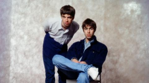 Noel Gallagher and Liam Gallagher from the band, Oasis, are in front of a beige background at a photoshoot in a hotel in Tokyo, September 1994. Noel is standing on the left and has brown hair, blue eyes, wears a white shirt, dark blue trousers, and is leaning over slightly. Liam is on the right, and has brown hair, blue eyes, wears a blue shirt, a dark blue jacket, blue jeans, white trainers, and is sitting down on a chair.