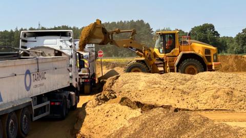A yellow digger on the right is dropping sand into a large which truck to the left, surrounded by a sandy quarry.