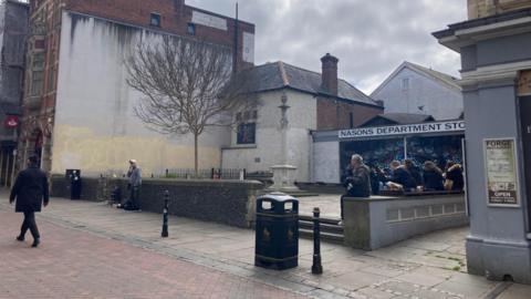 A small public square is surrounded by buildings on three sides. A public path runs along the square, and there are steps leading up to the square which has a war memorial and the entrance to the former Nason's department store. The main street is pedestrianised and there are bollards and a waste bin and people are walking around and sitting on the walls.