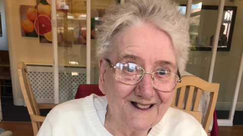 A woman with short grey hair, metal rimmed glasses and a cream top smiles at the camera.