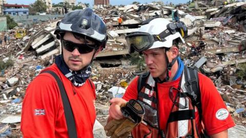 Rob Wingar is talking to a fellow volunteer in front of a huge pile of rubble. Both are wearing orange and protective helmets.
