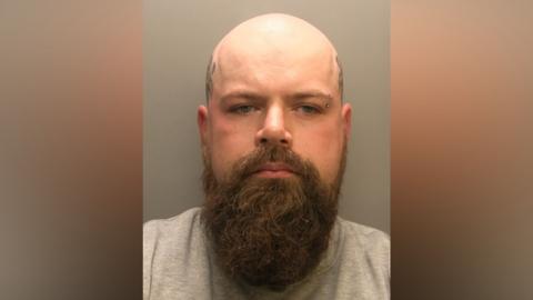 Police custody image of Courtney Gaston-Hird. He is bald and has a long brown beard. He is wearing a grey top and looking directly into the camera.