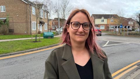 Alex Fisher standing by the side of the road near a school. She has red shoulder-length hair and wears glasses, a green coat and black top