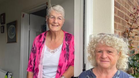 Sally Kidd (left) and Marion Parker (right). The pair are stood in a doorway with Kidd wearing a pink jacket and Parker is wearing a blue floral top.