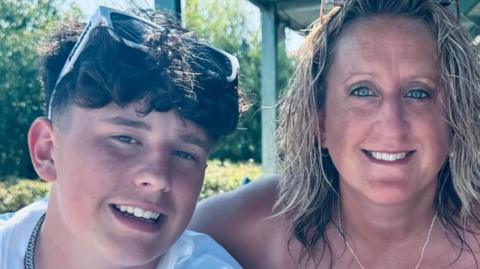 Alfie (who has dark hair and sunglasses on his head) and his mum (who has blonde hair) smile at the camera 