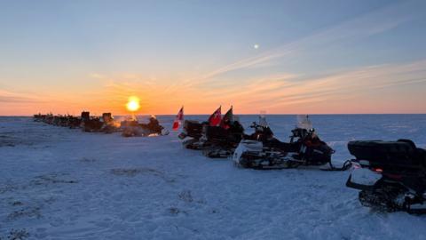 The patrol snowmobiles lines up on the border between the territory of Nunavut and the province of Manitoba. The sun is seen setting across the flat snow-covered terrain.