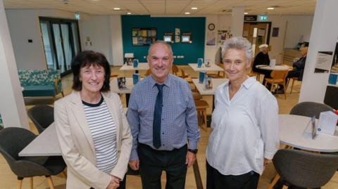 Amanda Healy, Durham County Council’s Director of Public Health, Councillor Howard Brown, Cabinet member for adult and health services, and Michelle Cooper, chief executive at Point North, smiling at the camera. There are tables and chairs in the room behind them. Amanda Healy has short brown hair and is wearing a beige suit jacket and a black and white striped top. Howard Brown has grey hair and is wearing a blue checkered shirt, black trousers and a blue tie. Michelle Cooper has short grey hair and is wearing black trousers and a white shirt with thin grey stripes. 

