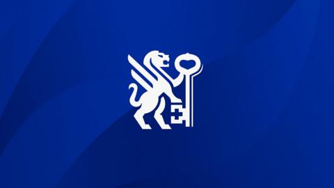 A football shirt crest shows a white winged lion standing up with a front leg on a key. It is white against a dark blue background