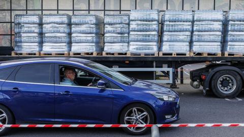 A man driving a blue car. The vehicle is stopped next to an open lorry trailer, which contains pallets of bottled water.