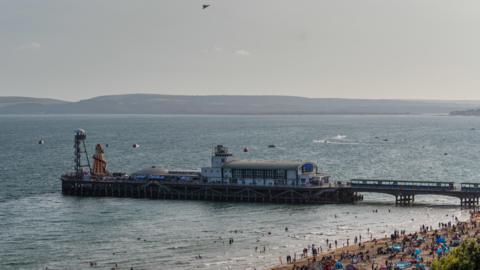 An aerial shot of Bournemouth Pier taken during the day. It comprised several buildings and structures, including a traditional helter-skelter slide. To the right, the edge of the beach is visible, with dozens of people gathered around by the sea. 