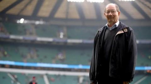 Ian Robertson standing in an empty sports stadium. He is wearing a black coat.
