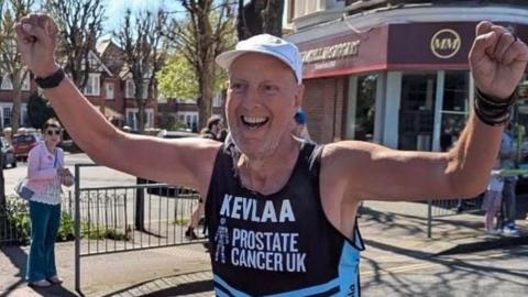 A close-up outdoor image of a runner on a pavement near a road junction, wearing a black “Prostate Cancer UK” vest and a white cap. Both of his arms are raised, hands clenched. A shopfront with signage and residential buildings appear in the background.
