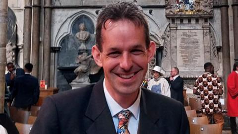Iain Farrington wearing a suit and a colourful tie smiling at the camera and standing in Westminster Abbey. There are chairs and people in the background as well as statues and an inscription on the wall.