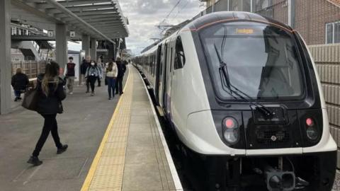 The front of an Elizabeth line train with a big glass front and white sides on the platform of an unidentified outdoor station with lots of people on the platform