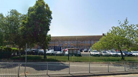 The entrance of Pensby High School with a blue sign on railings and the brown long building in the background, behind cars parked in the car park. 