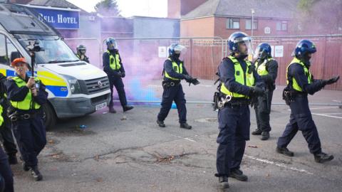 Several police officers in dark trousers, yellow high-visibility jackets and riot helmets stand outside The Bell Hotel in Epping. There is a police van on the left hand side of the photo, and a female police officer holding a camera on a monopod. There is blue and purple smoke in the background, and the hotel is blocked off by metal fencing.