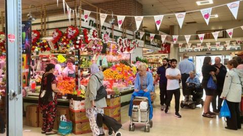 Lots of people standing around and shopping in an indoor market. One woman in a blue top pushes a shopping trolley while another with a lavender headscarf pulls her trolley. The stall they are near has bright oranges and flowers on display.