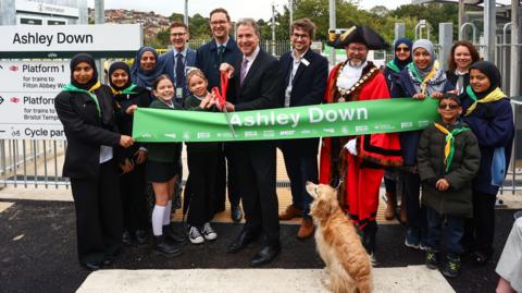 Various dignitaries, including the mayor of bristol, the mayor of the west of england Dan Norris and a group of school children holding a pair of giant scissors in front of a banner with the words "Ashley Down", preparing to cut the banner