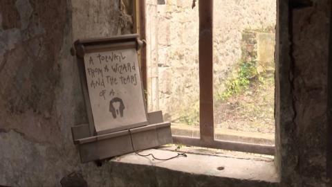 A scroll from the show leaning on a dusty windowsill in Gwyrch Caste. It says 'A toenail from a wizard and the tears of a ...'. Below the text is an image of an outline of a mummy's head. 