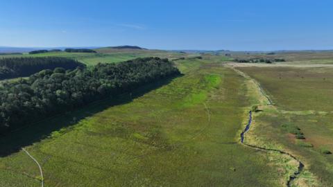 An aerial photograph of the land which has a slim river meandering across the green open land. There is a forest to the left and hills can be seen in the distance.