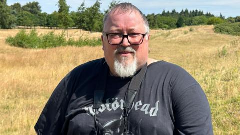 A man with short grey hair, a white goatee and black-rimmed glasses standing on a patch of grassland on a sunny day. Many trees are in the distance. He is wearing a black Motorhead t-shirt and has a pair of binoculars hanging around his neck.